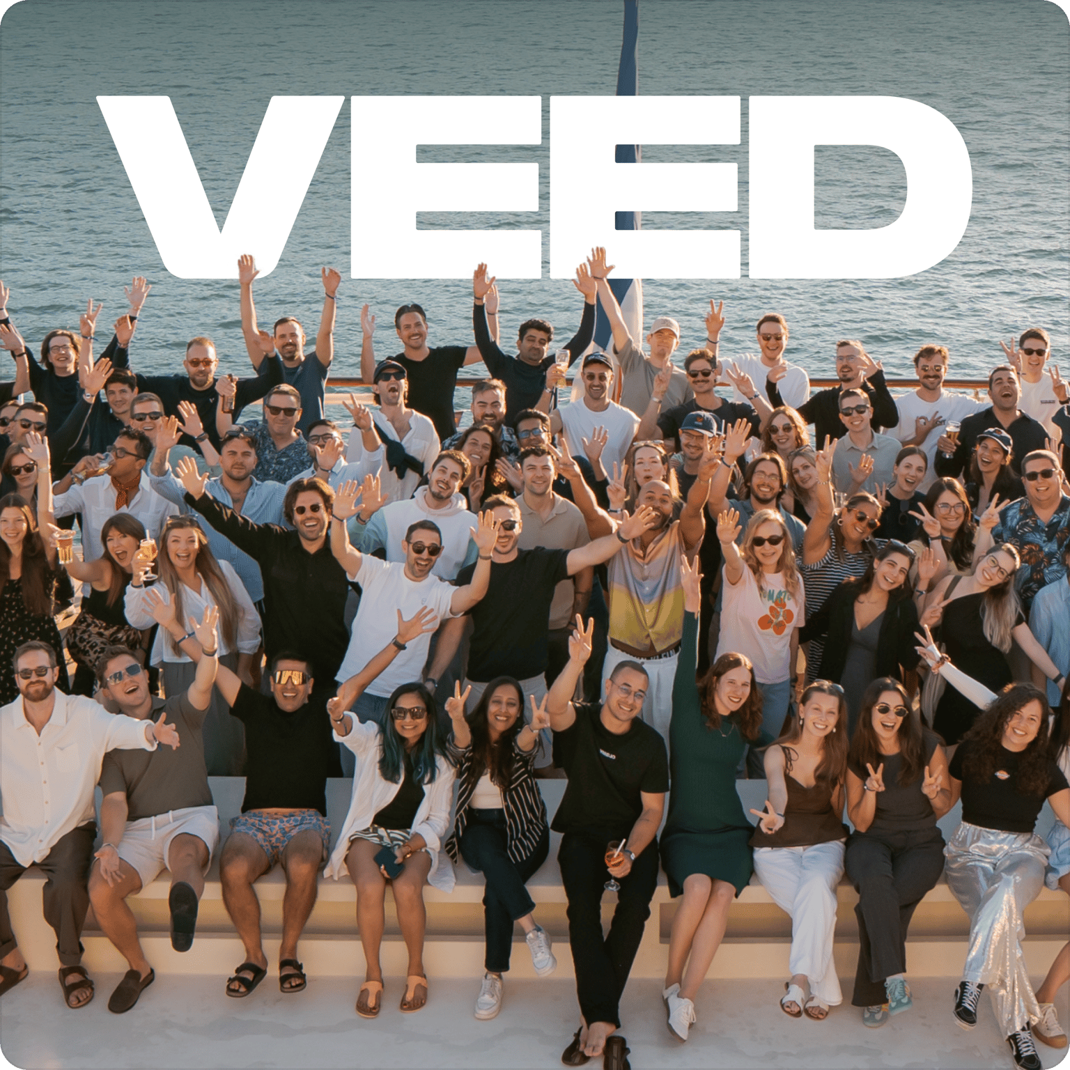 VEED About Us header showing a large team of employees smiling and waving together outdoors by the water