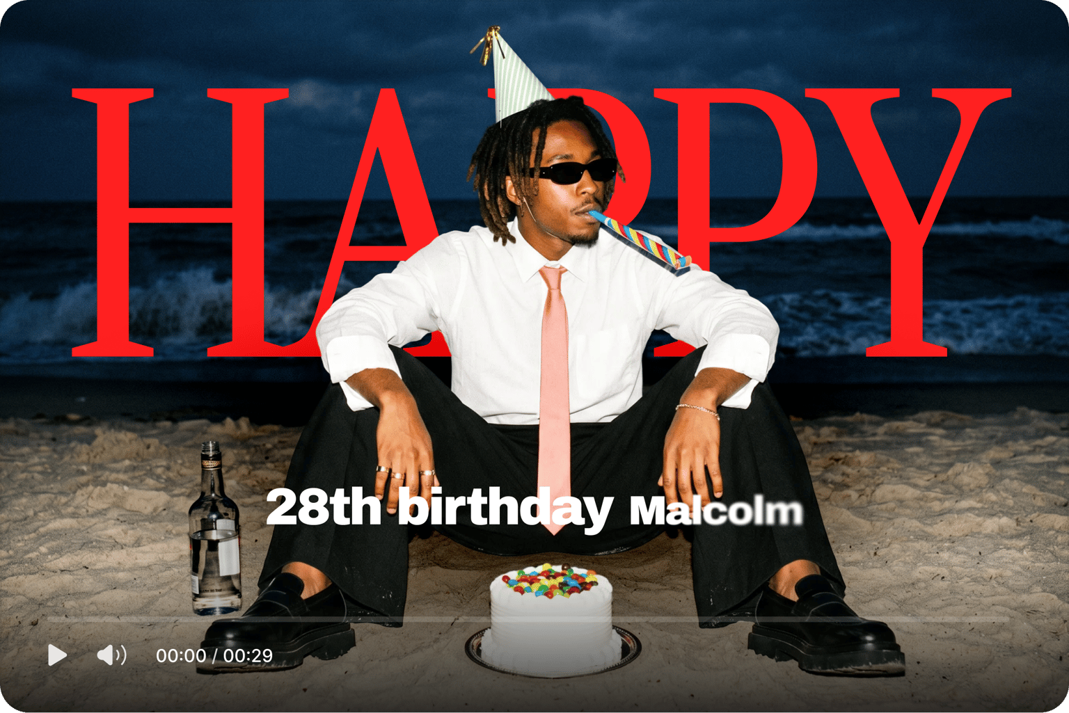 Birthday video maker showing happy 28th birthday Malcolm text over a man in a party hat sitting on a beach with a cake