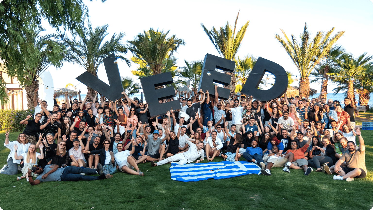 A large diverse group of people celebrating and cheering around large black letters spelling VEED on a grassy field in a beach resort setting.