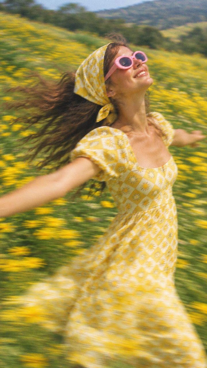 A woman in a yellow sun-print dress and pink sunglasses spinning in a field of yellow flowers